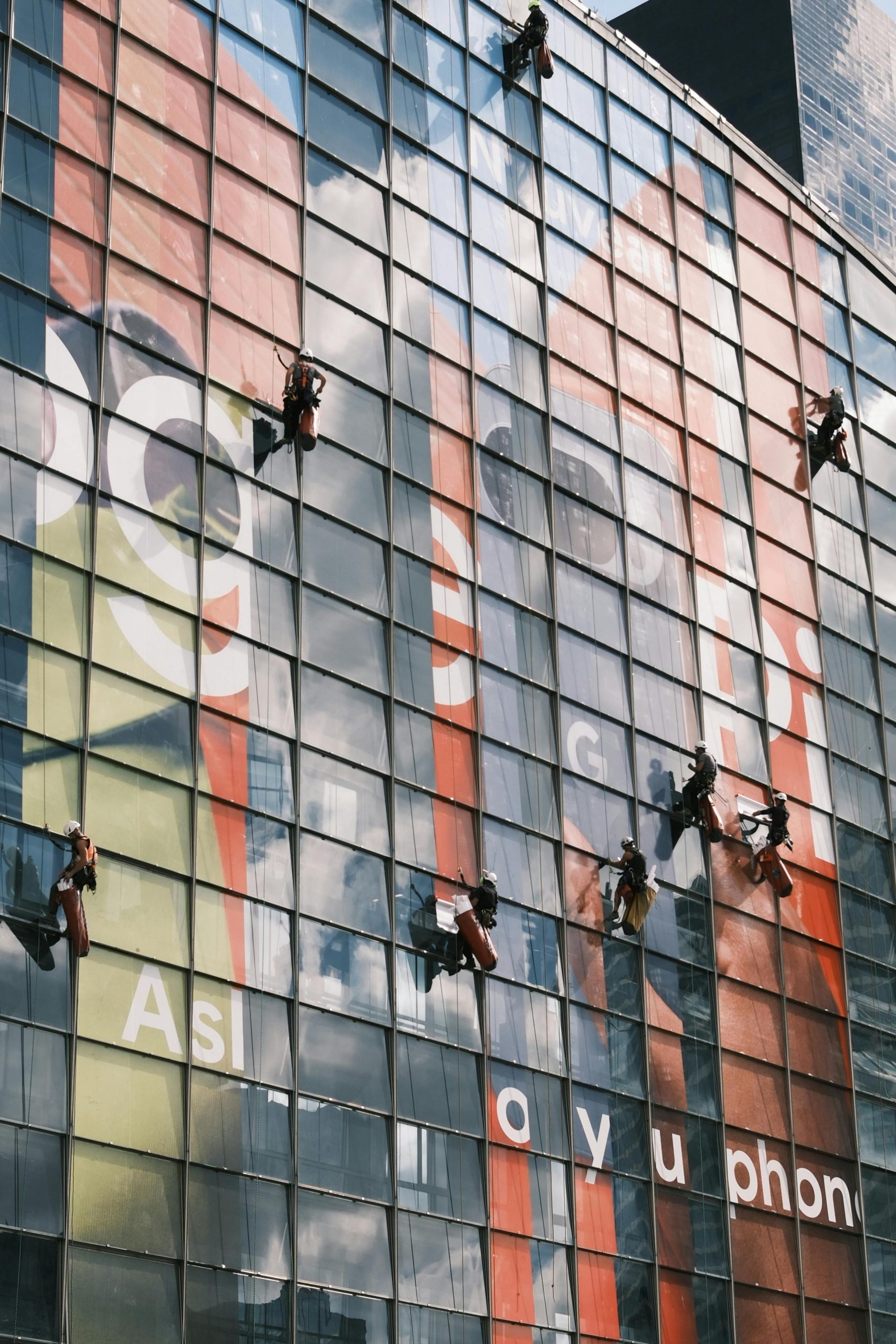 Workers scale the facade of a high-rise building to clean glass panels with visible advertisements.